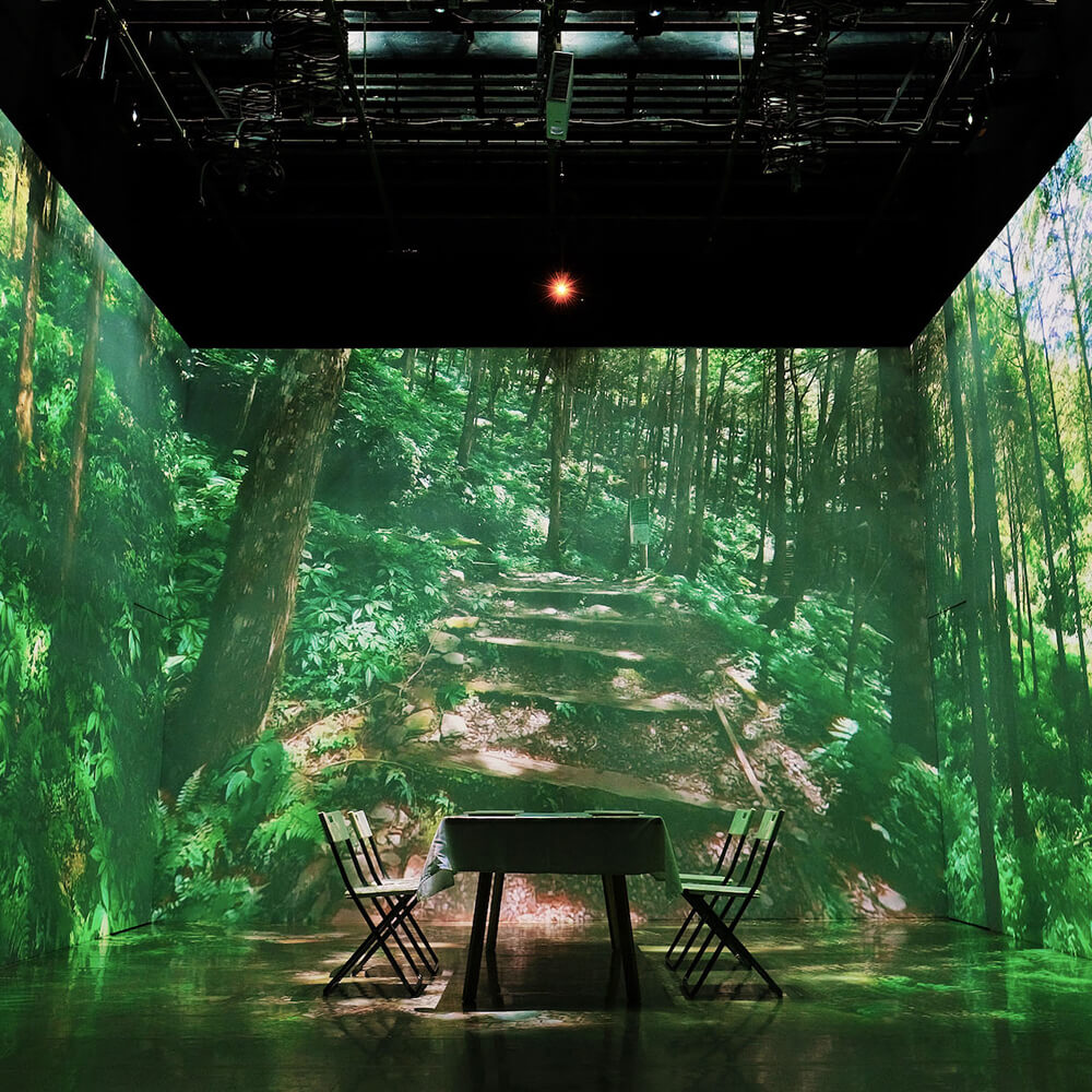 A tranquil forest scene is projected onto the walls and ceiling of an intimate dining space, creating a serene woodland atmosphere. A small table and chairs sit surrounded by lush green foliage and dappled sunlight, offering diners an escape into nature.